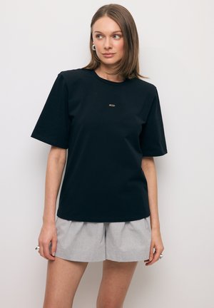 Kobieta ubrana w czarną koszulkę z krótkim rękawem z małym złotym logo oraz jasnoszare plisowane szorty na tle białego, jednolitego tła.