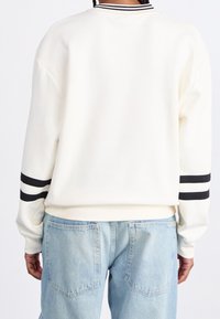 Felpa bianca con strisce nere sulle maniche, colletto e polsini a coste. Indossata con jeans in denim blu chiaro, che mostrano una vestibilità ampia.