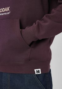 Personne portant un sweat à capuche Kodak violet foncé avec une main dans la poche, associé à un jean en denim bleu.