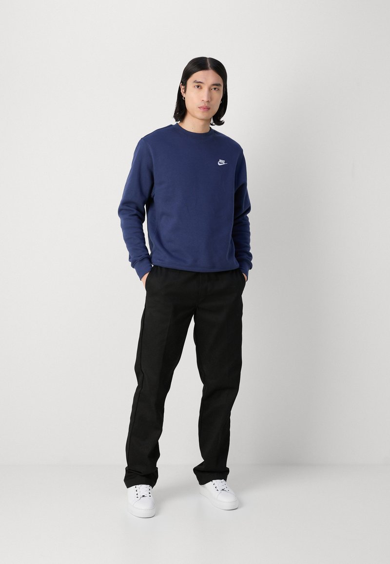 Marinblå sweatshirt med rund hals och liten vit logotyp, i kombination med svarta byxor och vita sneakers. Enkel, avslappnad passform.