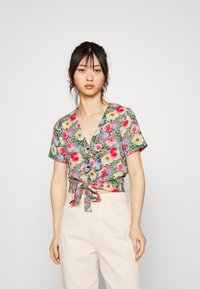 Blusa floral de mangas curtas com decote em V, detalhe de laço na frente e padrões vibrantes de rosa, amarelo e verde sobre um fundo creme. Combinada com calças claras.