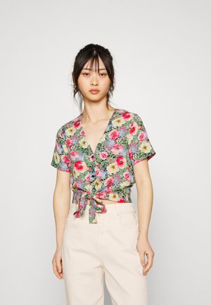 Blusa floral de mangas curtas com decote em V, detalhe de laço na frente e padrões vibrantes de rosa, amarelo e verde sobre um fundo creme. Combinada com calças claras.