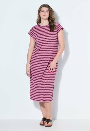 Kobieta stojąca, ubrana w różową sukienkę do kolan z czarnymi okrągłymi wzorami oraz czarne sandały z odkrytymi palcami na białym tle.