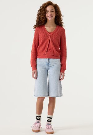 Cárdigan rojo de punto sobre una camiseta a juego, combinado con culottes de mezclilla azul claro. Zapatillas deportivas rosas y calcetines a rayas en blanco y negro completan el conjunto.