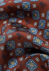 Tela marrón con patrones florales en azul, blanco y amarillo, así como pequeños diseños de calaveras y tibias, mostrando textura y pliegues.