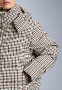 Padded kaput s visokim ovratnikom, s uzorkom houndstooth u nijansama kreme, burgunde, plave i crne, s bočnim džepovima.