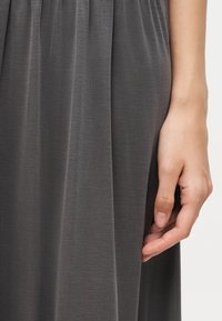 Το δεξί χέρι ακουμπά δίπλα σε ένα γκρι, πλισέ φόρεμα από ύφασμα σε ουδέτερο φόντο.