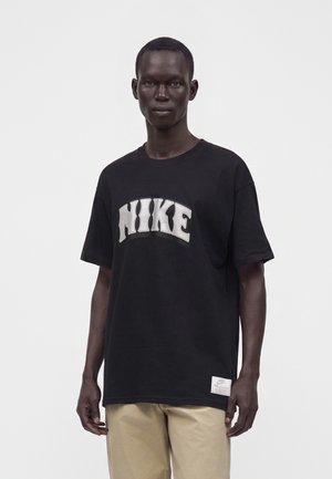 Homme portant un t-shirt noir Nike avec un grand logo blanc et un pantalon beige, debout devant un fond blanc uni.