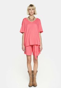 Różowa bawełniana koszulka z krótkim rękawem, o warstwowej konstrukcji, zestawiona z pasującymi różowymi szortami i brązowymi kowbojkami, na białym tle.