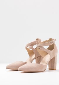 Beiga mocka högklackade skor med spetsig tå, ankelsnören och tjock klack. Designen har korsade remmar och en slät textur.