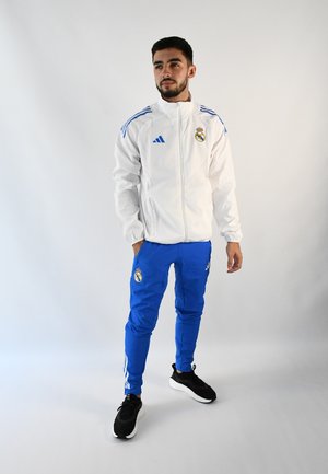 Chaqueta blanca con cremallera y detalles en azul, que presenta el logotipo del Real Madrid. Unos pantalones deportivos azules y unas zapatillas deportivas negras completan el conjunto.
