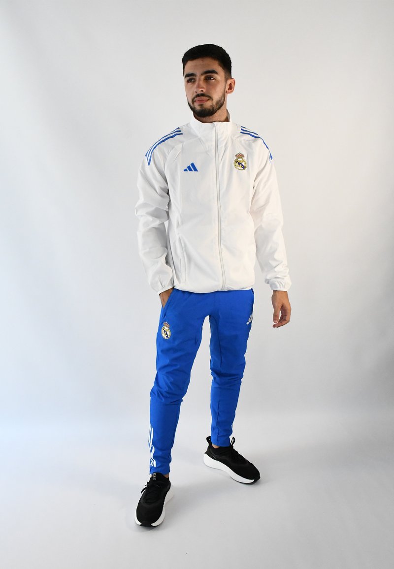 Chaqueta blanca con cremallera y detalles en azul, que presenta el logotipo del Real Madrid. Unos pantalones deportivos azules y unas zapatillas deportivas negras completan el conjunto.