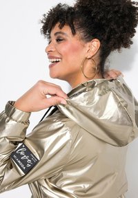 Metalen gouden hooded jack met elastische manchetten en een patch met de tekst "Gemaakt met liefde door Miamoda" op de linkermouw.