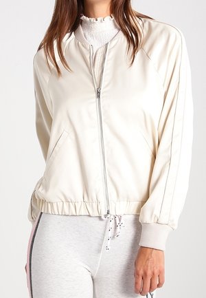 Kvinde iført en lys beige bomberjakke med lynlås over en hvid bluse med flæser, kombineret med lysegrå leggings med sidelommer.