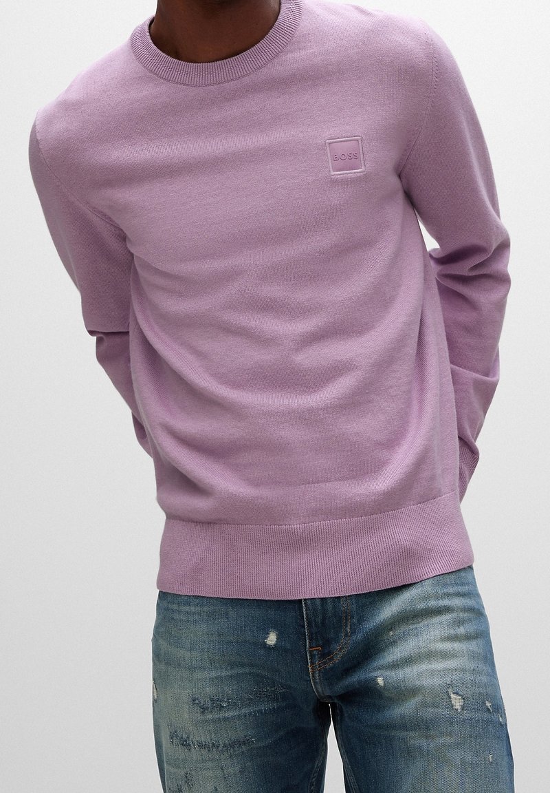 Homme portant un pull violet clair à col rond avec un petit logo "BOSS" et un jean bleu délavé sur un fond uni.