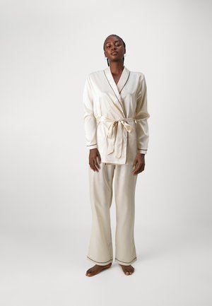 Ivory satin pajama set with a wrap jacket featuring black piping, long sleeves, and matching wide-leg pants. Features a tie belt and pockets.