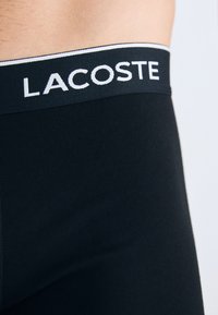 Close-up van de zwarte onderbroekenband met het witte Lacoste-logo, gedragen om de taille van een persoon tegen een effen achtergrond.