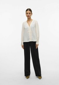 Femme portant un blouse crème à col en v avec des boutons, un pantalon noir à jambes larges et des chaussures pointues olivées, se tenant devant un fond blanc uni.