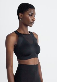 Mujer con cabello corto, vistiendo una camiseta sin mangas negra y leggings de cintura alta, de pie contra un fondo blanco liso.
