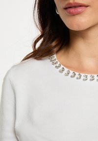 Jersey de punto blanco con cuello redondo adornado con embellishments decorativos plateados. Textura suave con un diseño ajustado.
