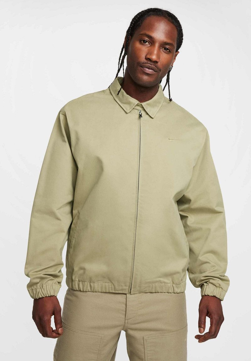 Chaqueta de manga larga en verde olivo claro, con cuello tipo camisa, puños elásticos, cremallera frontal y un diseño minimalista, con un discreto detalle del logo.