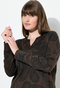 Top marrón de manga larga con un patrón de paisley en tonos oscuros. Presenta un escote en V y una tela texturizada. El modelo posa con las manos entrelazadas.