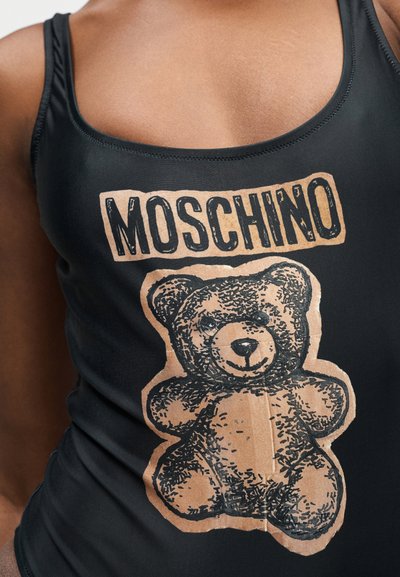 Μαύρο αμάνικο μπλουζάκι με γραφικό καφέ αρκουδάκι και τη λέξη "MOSCHINO" τυπωμένη πάνω στο άνω μέρος του κορμού ενός ατόμου.