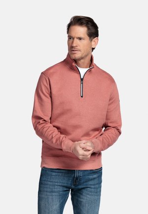 Man met een zalmkleurige sweatshirt met een kwart-ritssluiting over een wit overhemd, met gevouwen handen, die iets opzij kijkt tegen een witte achtergrond.