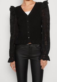 Svart cardigan med ribbat mönster, V-hals och knäppning fram. Långa ärmar med broderade blommönster och volangärmar.