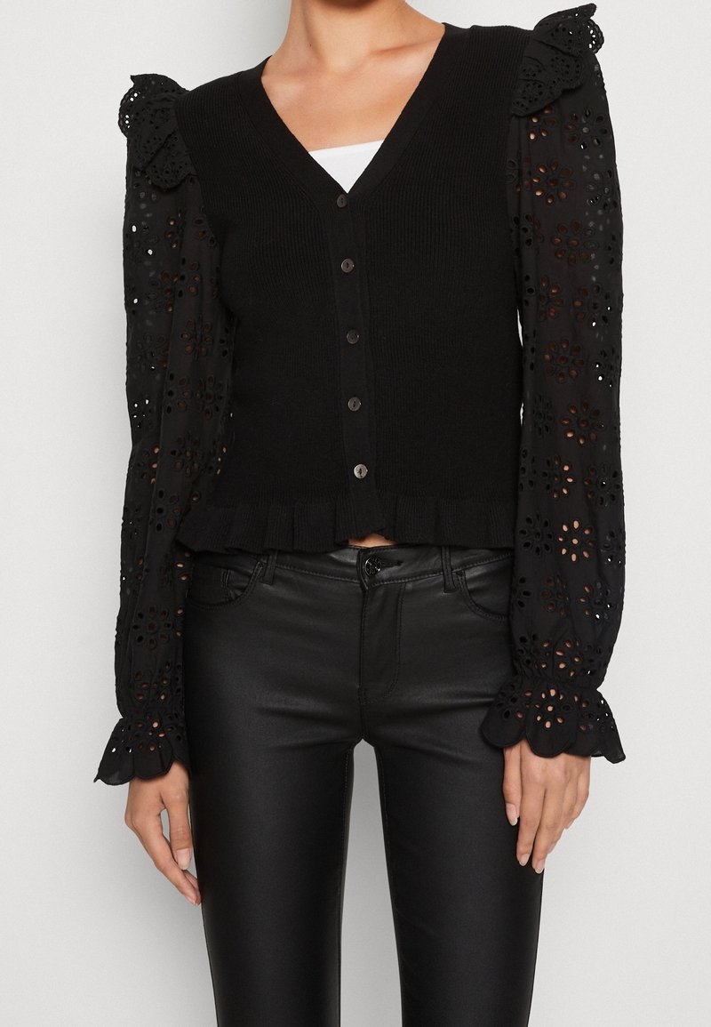 Svart cardigan med ribbat mönster, V-hals och knäppning fram. Långa ärmar med broderade blommönster och volangärmar.