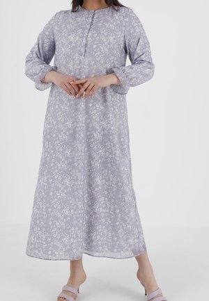 Nainen, jolla on pitkä, vaalean violetti kukkakuvioinen mekko pitkine hihoineen, seisoo paljain jaloin valkoista taustaa vasten.