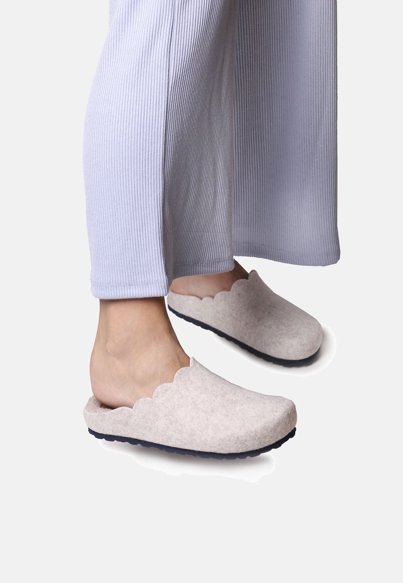 Gaišpelēku slip-on apavi ar viļņotu malu un teksturētu virsmu, redzami uz kājām, kas ģērbtas gaismas violetos ribota auduma biksēs.