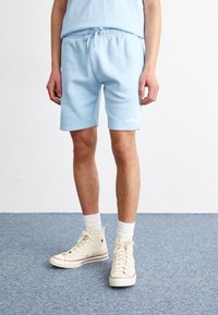 Personne portant un short à cordon de serrage bleu clair, des chaussettes blanches montantes et des baskets montantes beige pâle, debout sur un tapis bleu.