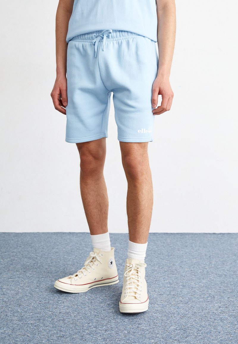 Personne portant un short à cordon de serrage bleu clair, des chaussettes blanches montantes et des baskets montantes beige pâle, debout sur un tapis bleu.