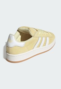 Zapatillas de ante amarillas con acentos blancos, tres franjas blancas, un collar acolchado y una suela de goma texturizada. Cierre de cordones ajustable.