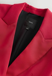 Rød blazer med et glat, struktureret stof, der har en skarp krave og en sort indvendig foring. Inkluderer et "NEXT"-mærke.