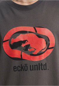 Grå bomull t-skjorte med et stort rødt og svart grafisk motiv av et stilisert dyr innenfor en oval form, med "eckō unltd." tekst under.