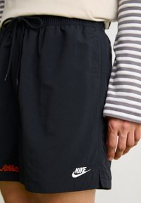 Person med svarta Nike-shorts med broderad logotyp, tillsammans med en långärmad randig skjorta, visar handen vilande bredvid shortsen.