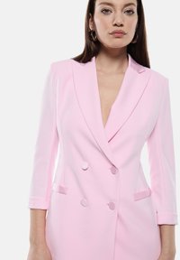 Blazer rosa doppiopetto con scollo a V profondo, spalle strutturate e dettagli con bottoni. Tessuto morbido e vestibilità su misura.