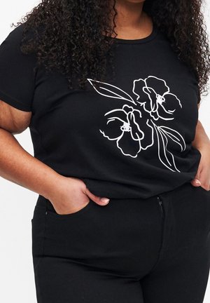 Vrouw met krullend haar, gekleed in een zwarte broek en een zwart T-shirt met een witte, omlijnde bloemmotief, handen deels in de zakken.