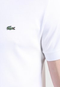 Bela polo majica z rebrastim ovratnikom in manšeti. Ima majhen zeleni krokodilov logotip vezen na prsih. Gladka, bombažna mešanica tkanine.