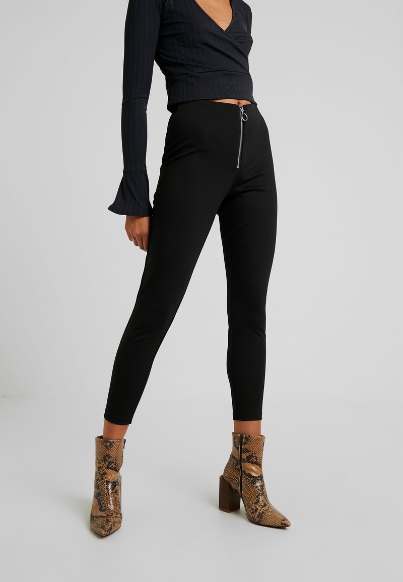 Femme portant des leggings noirs taille haute avec fermeture éclair devant, un crop top noir à manches longues et des bottines marron en cuir façon peau de serpent avec talons blocs.