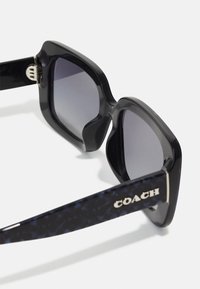 Juodos saulės akiniai su gradientiniais lęšiais, plačiomis rėmeliais ir tekstūruotais mėlynais raštais ant skruostų. Šone yra sidabrinis "COACH" logotipas.