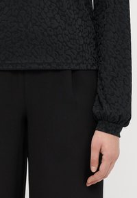 Top preto texturizado com um discreto padrão de animal print, apresentando mangas longas e volumosas. Combinado com calças pretas ajustadas.