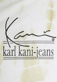 White fabric with a tie-dye pattern in light yellow, featuring the text "Karl Kani" in stylized black font above "karl kani-jeans."