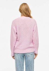 Femme aux longs cheveux blonds portant un pull rose à maille ouverte et un jean bleu clair, vue de dos sur un fond blanc uni.