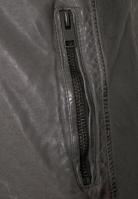 Grau Lederjacke mit einer seitlichen Reißverschlusstasche aus schwarzem Metall und glatter Textur, die die strukturierten Designelemente hervorhebt.