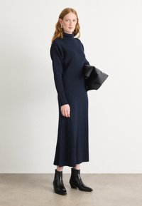 Vestido de punto acanalado en azul marino con cuello alto y mangas largas, combinado con botas negras de tobillo. La modelo sostiene un bolso de mano negro.