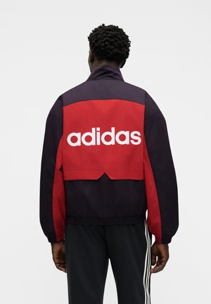 Muž zády, oblečený v černo-červené bundě s bílým nápisem „adidas“ na zádech, k tomu černé kalhoty s bílými pruhy.