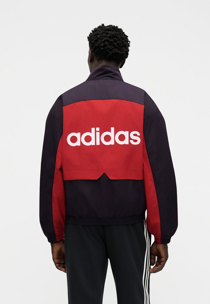 Muž zády, oblečený v černo-červené bundě s bílým nápisem „adidas“ na zádech, k tomu černé kalhoty s bílými pruhy.